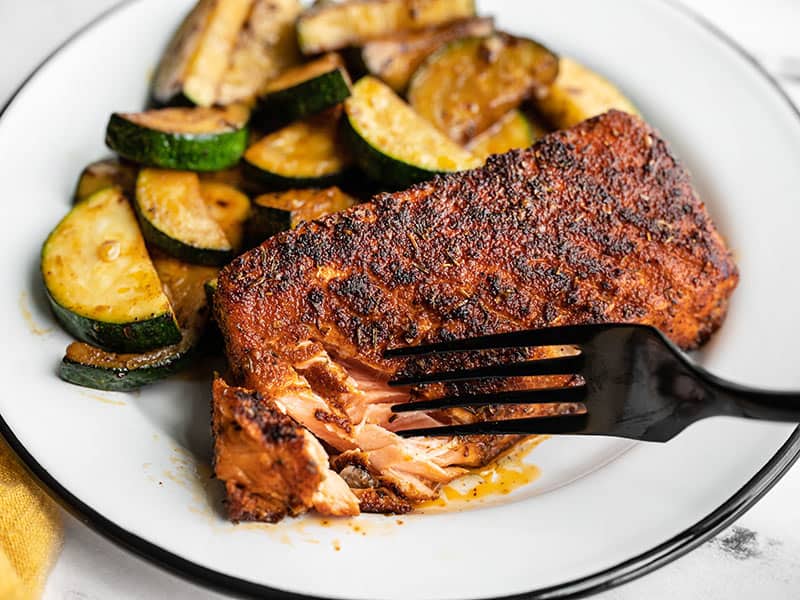 Blackened salmon coated in Cajun spices, cooked to perfection in butter, and paired with simple sautéed zucchini makes a simple but delicious dinner! Budgetbytes.com A fork flaking a piece of blackened salmon on the plate with zucchini