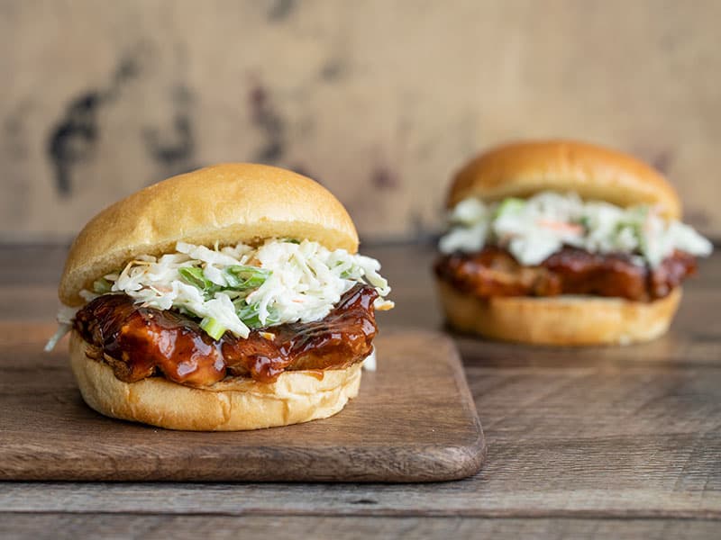 These Easy BBQ Chicken Sandwiches are a fast, easy, and delicious way to satisfy your BBQ craving. No grill needed! Perfect for busy weeknights. Budgetbytes.com Two BBQ Chicken Sandwiches, one on a cutting board, the other in the background.
