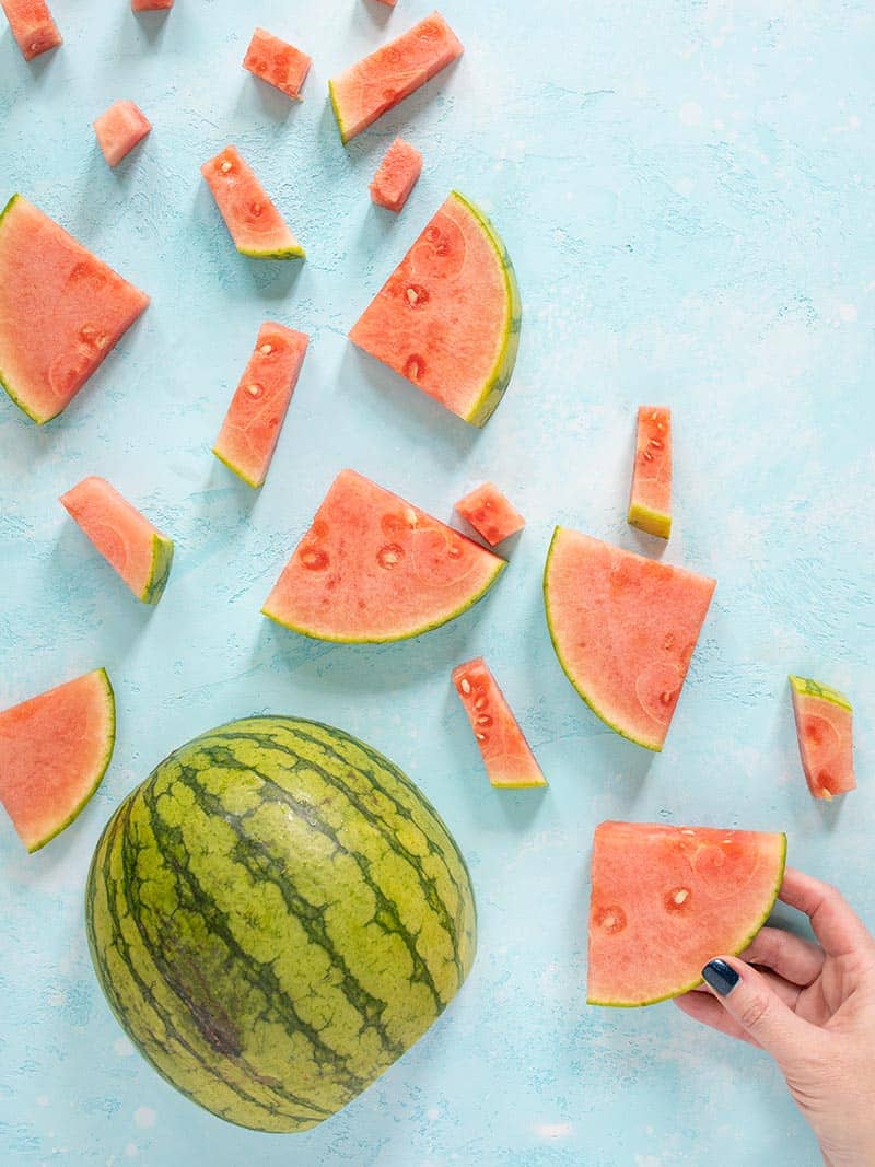 A step by step guide on how to cut watermelon into classic wedges or easy-to-eat batons! Make your summer easy and delicious! Budgetbytes.com A watermelon cut into several different shape and sized pieces, a hand grabbing a wedge