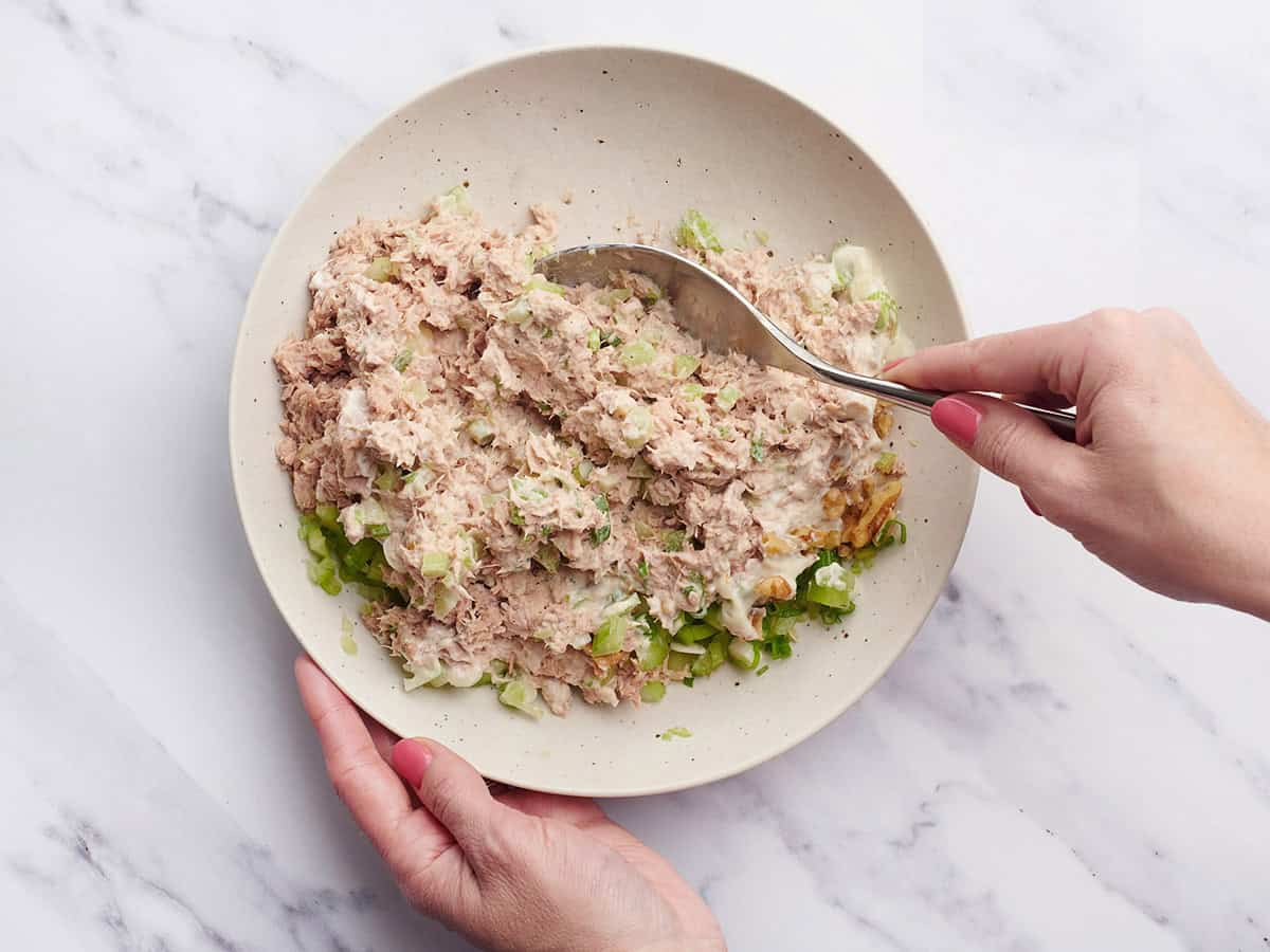 A spoon mixing together the ingredients for tuna salad in a bowl.