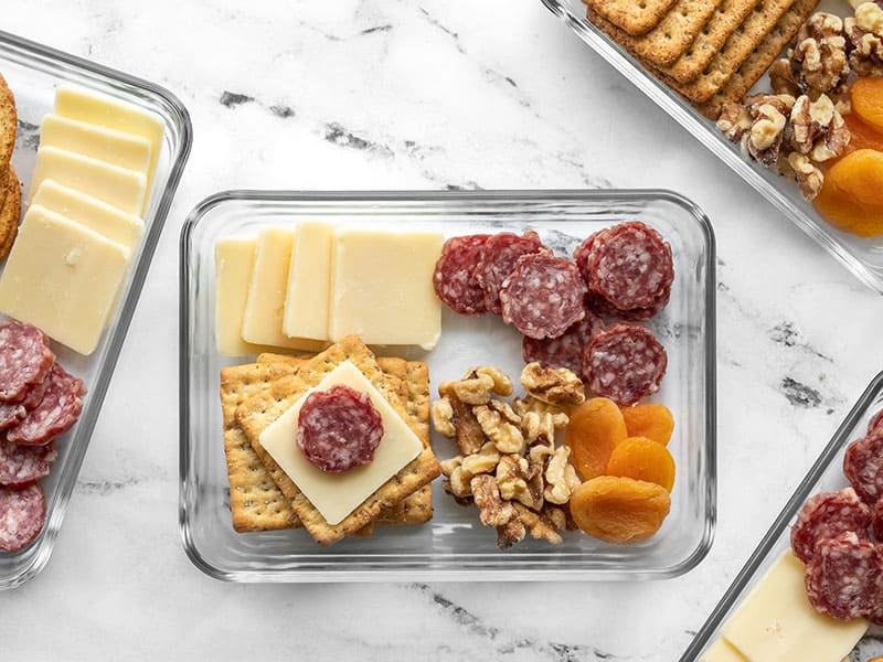 This easy no-cook lunch idea is perfect for the cheese lovers of the world! The Cheese Board Lunch Box makes a meal out of savory nibbles. Budgetbytes.com Overhead view of one Cheese board Lunch Box with a cracker stacked with cheese and salami.