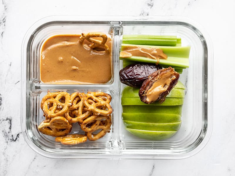 This peanut butter lunch box is an easy no-cook lunch idea for school, work, picnics, or road trips! No reheating needed! Budgetbytes.com A single peanut butter lunch box with peanut butter on a Medjool date and a pretzel dipping into the peanut butter