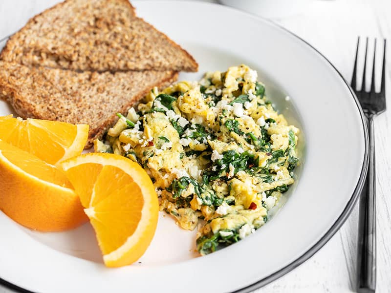 These tasty scrambled eggs with spinach and feta are fast and easy enough to prepare on a weekday, and are perfect for using up spinach! Budgetbytes.com Side view of a plate with scrambled eggs with spinach and feta, toast, and oranges