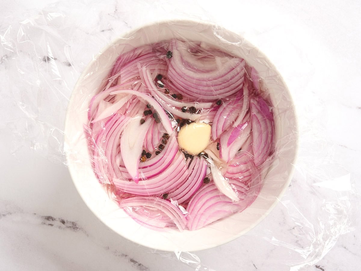 Plastic wrap placed over a bowl of sliced red onions, pepper corns and garlic.