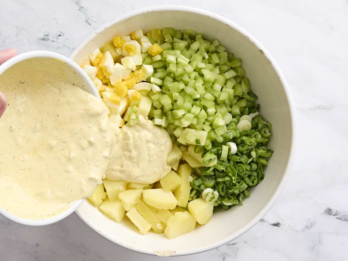 The dressing being poured into a bowl of Southern style potato salad ingredients.