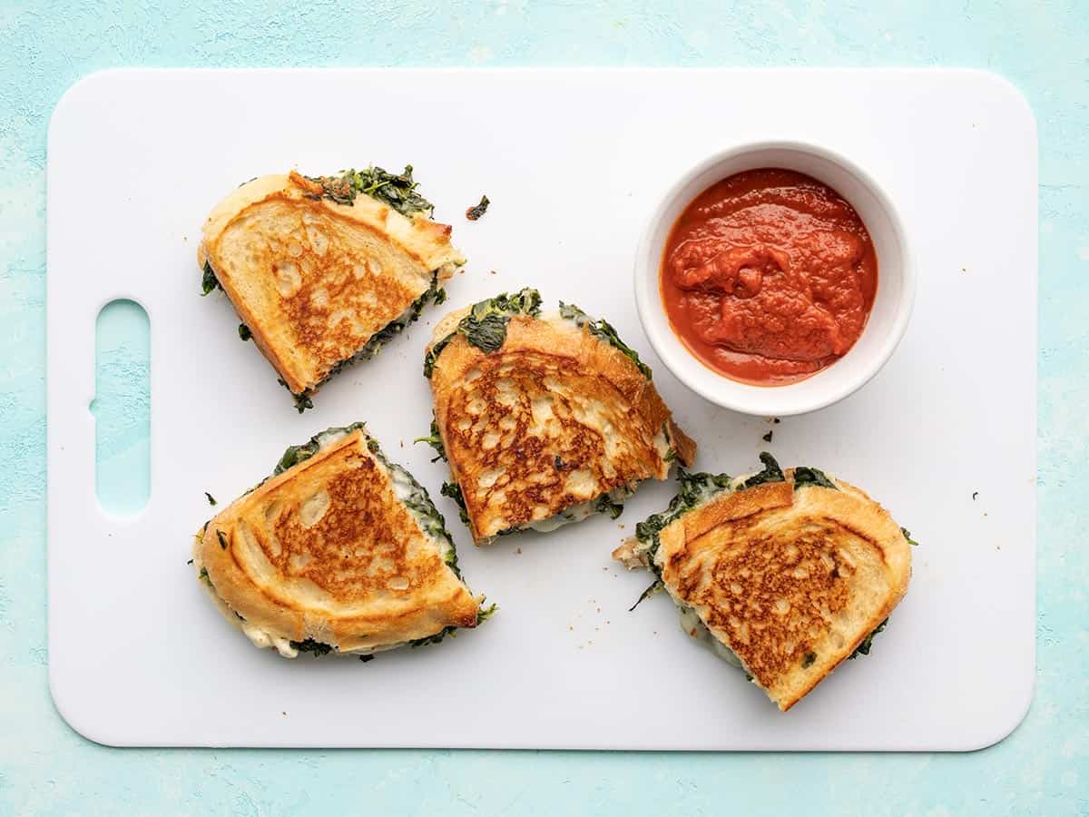 Overhead view of spinach and feta grilled cheese on a cutting board with a bowl of sauce.