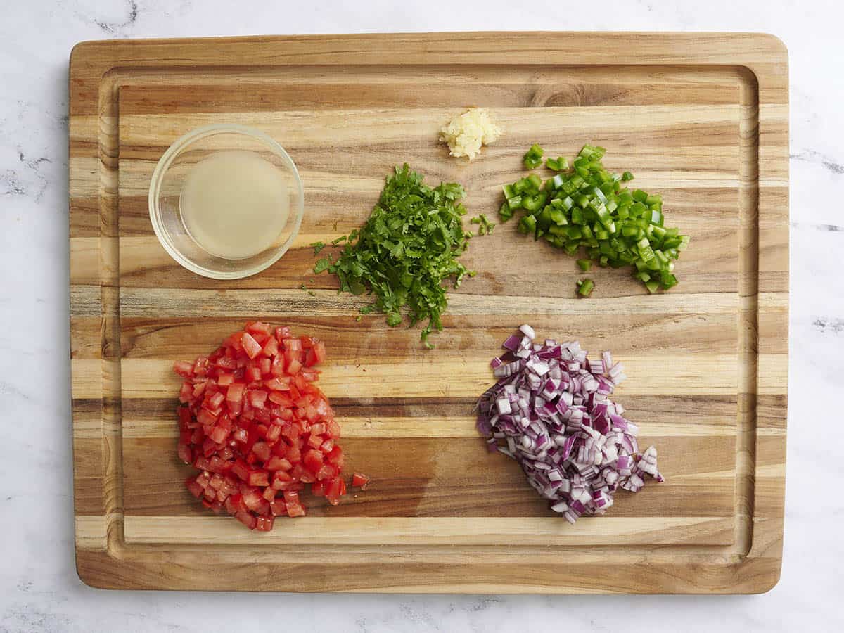 Diced tomato, cilantro, red onion, green bell pepper, minced garlic, and lemon juice on a wooden cutting board.