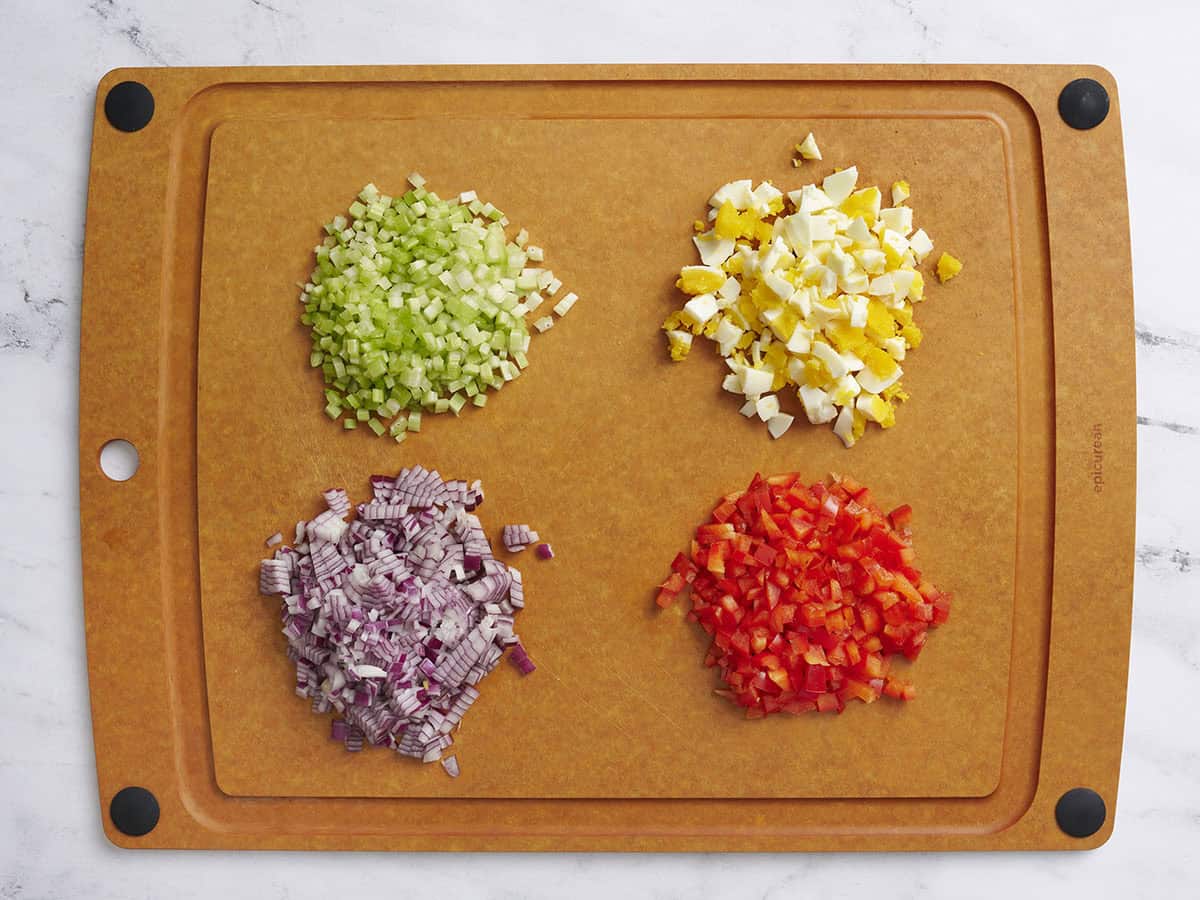 Diced celery, hard boiled egg, red onion, and red bell pepper on a wooden butting board.