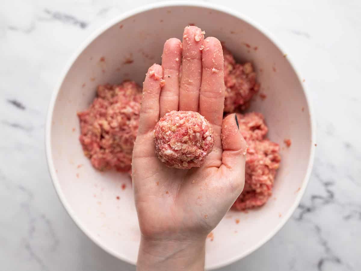 Shaped meatball held in a hand with a bowl of the meat mixture behind.