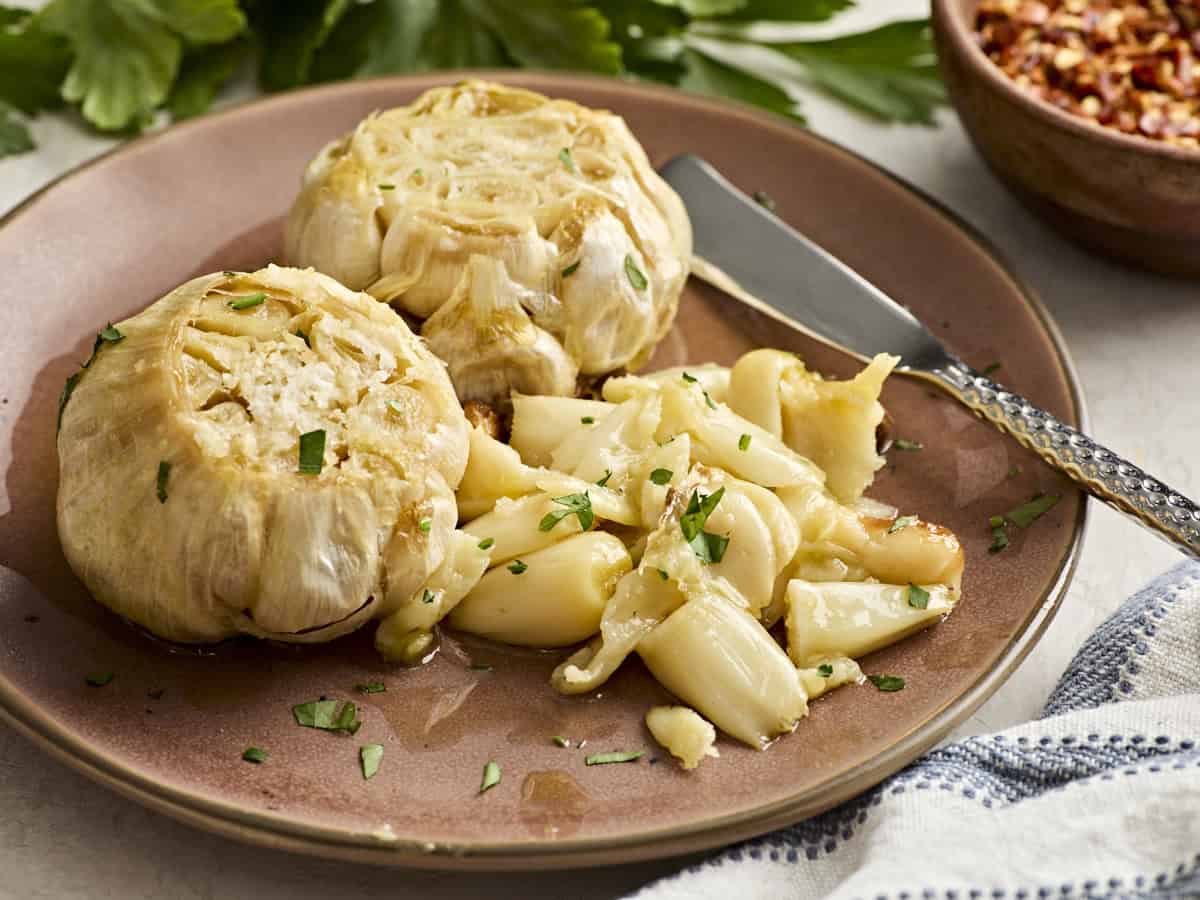 Side view of two head of roasted garlic on a plate with a knife and some garlic cloves.