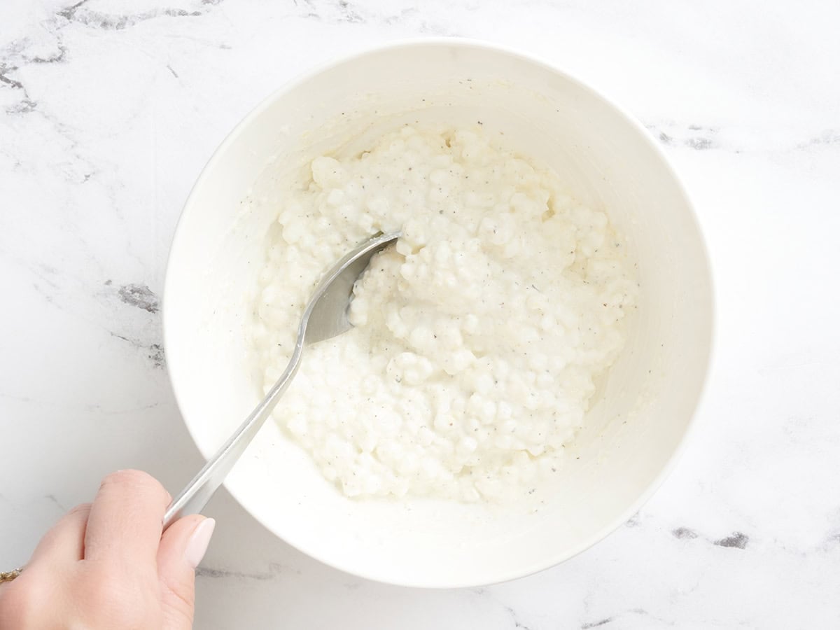 A spoon mixing together cottage cheese, parmesan, and black pepper in a bowl.
