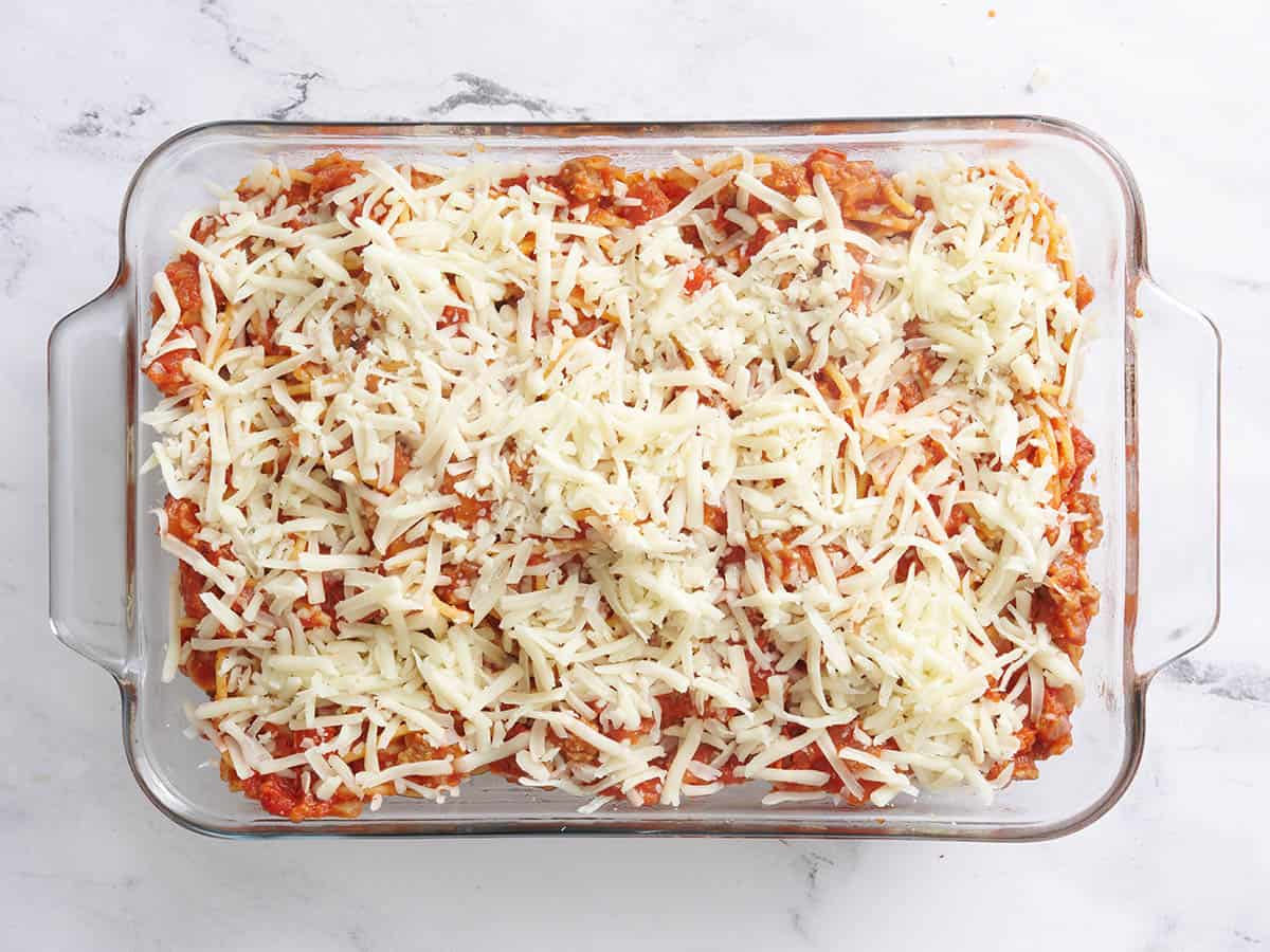 Shredded cheese sprinkled over baked spaghetti in a baking dish before baking.