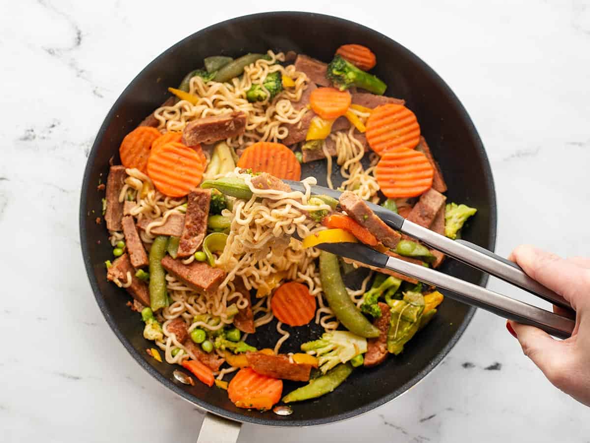 Spam stir fry noodles in a skillet, being lifted by tongs.