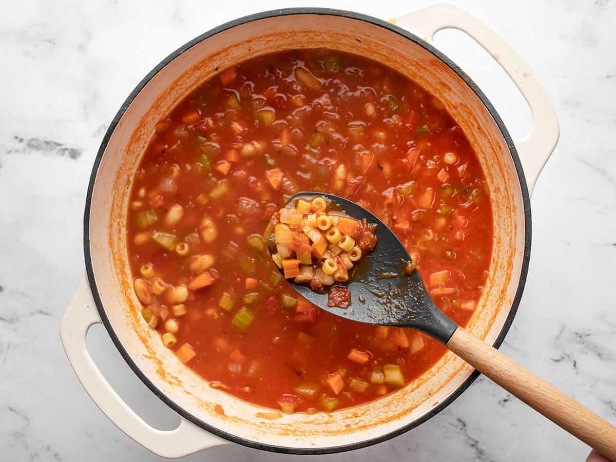 A close up shot of a wooden spoon raised above a cooked pot of pasta e fagioli soup.