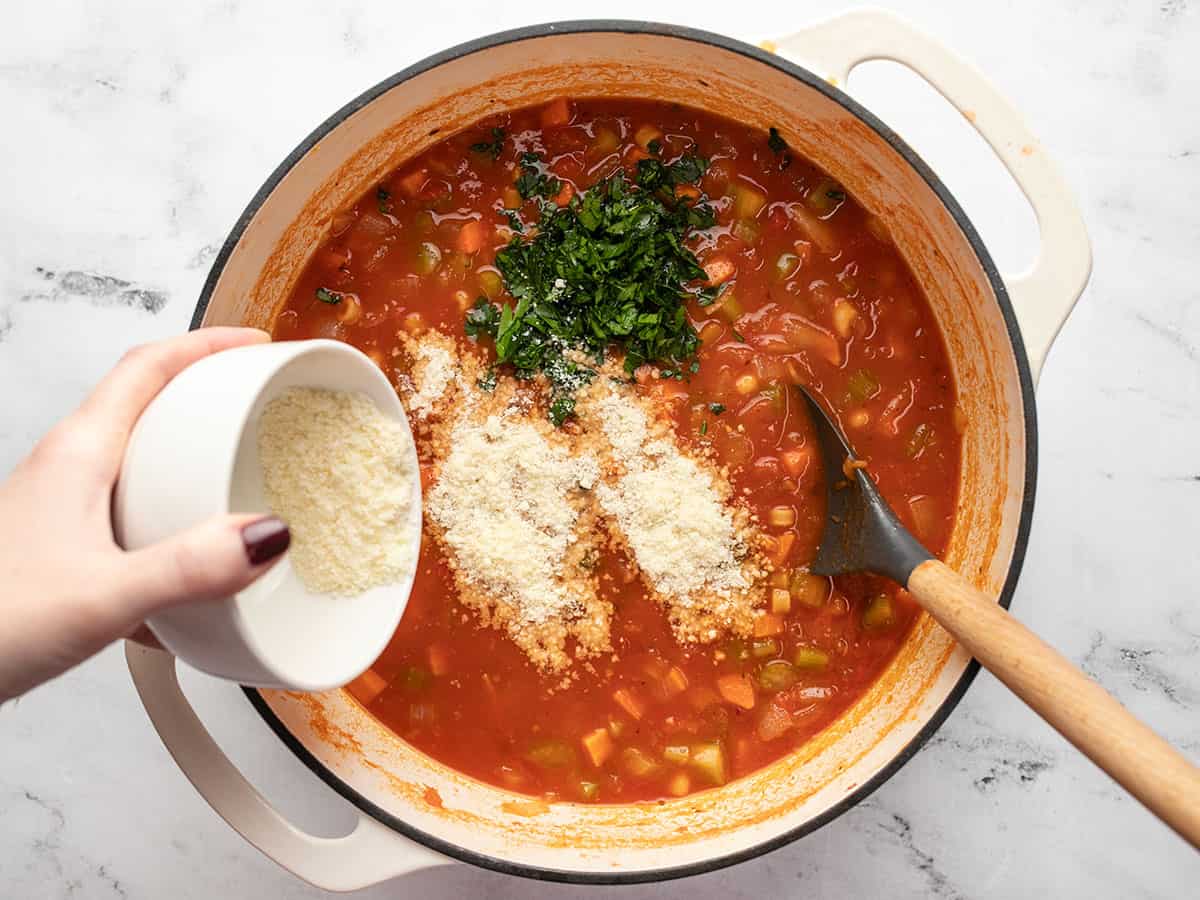 A large pot filled with cooked pasta e fagioli soup with a wooden spoon on the right side of the pot, and a hand tossing in grated parmesan cheese and chopped parsley sitting on top of the soup inside the pot.