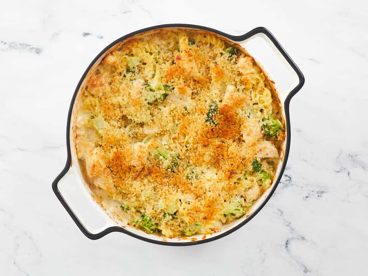 A chicken alfredo bake with broccoli topped with melted cheese and golden brown toasted breadcrumbs in a round, white enamel baking dish with a black rim that's sitting on a white, marble background.