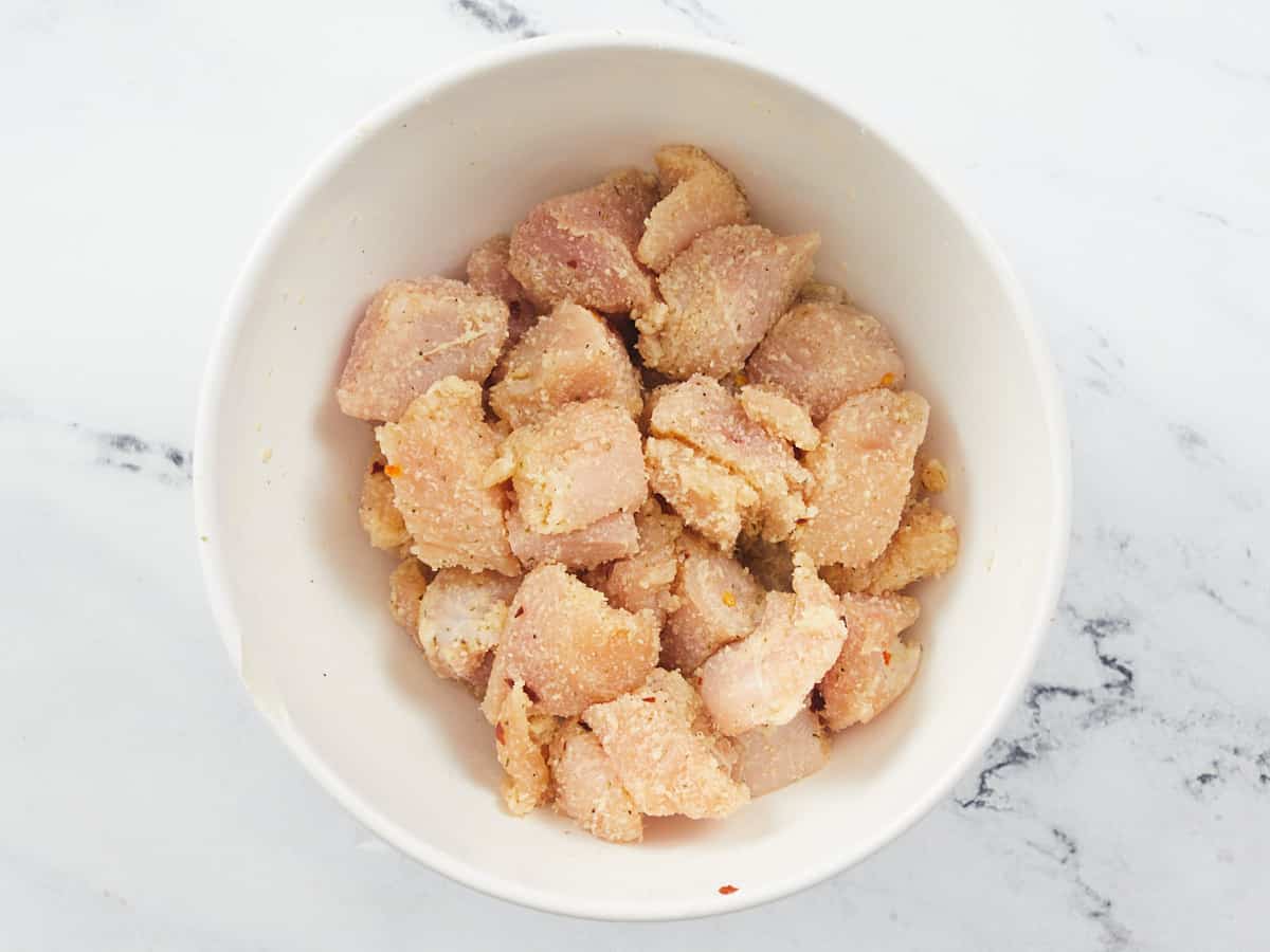 A white bowl of uncooked chicken breast chunks seasoned with grated Parmesan cheese, salt, black pepper, Italian seasoning and red pepper flakes. The bowl is sitting on a marble background.