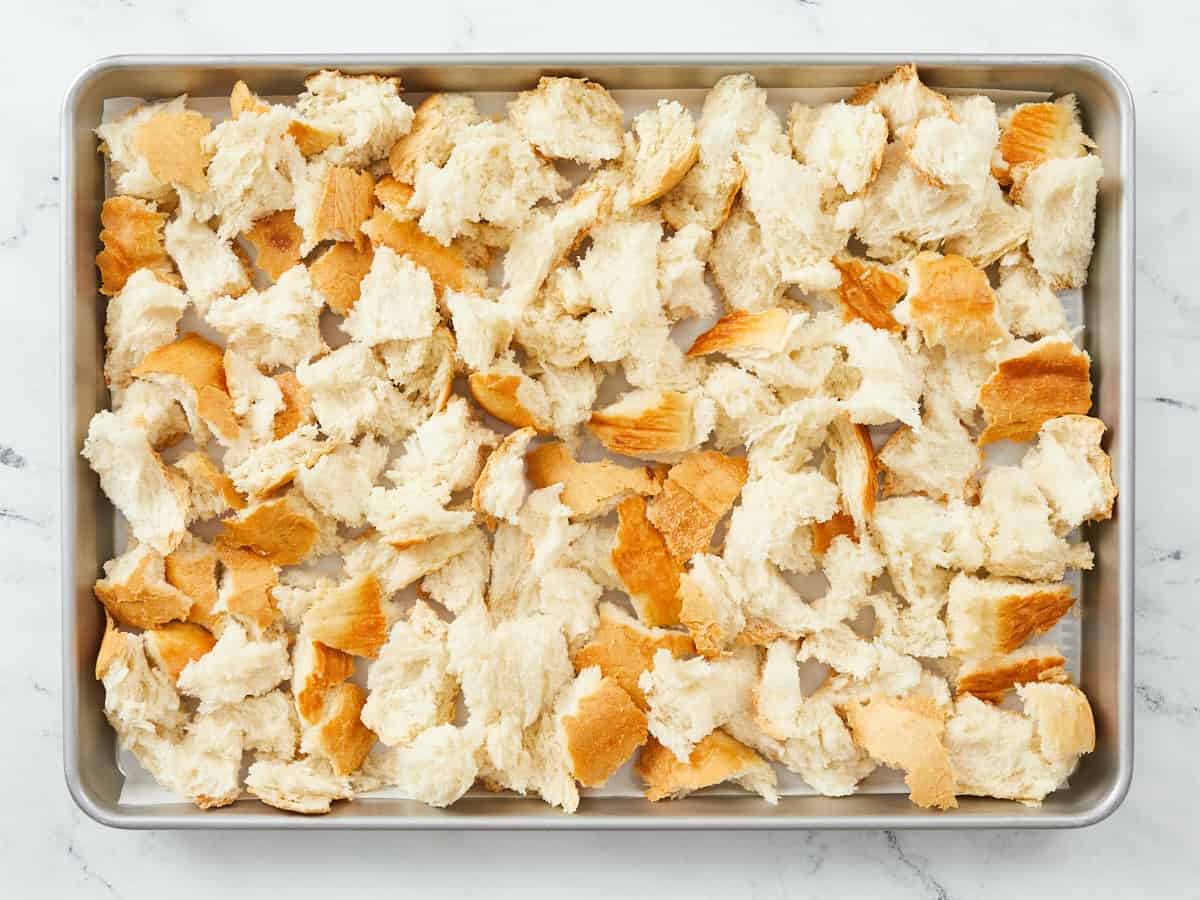 A rimmed metal baking sheet lined with parchment paper and covered with torn pieces of bread sitting drying out to make a bread pudding, and the sheet pan is sitting on a white marble background.