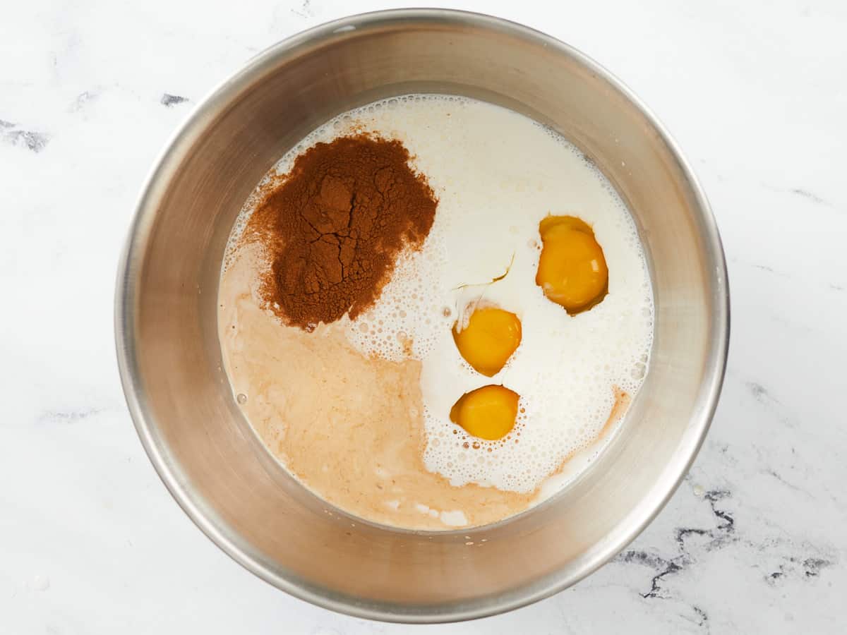A large metal mixing bowl filled with separated ingredients for bread pudding, which are eggs, half & half, vanilla extract, ⅔ cup of sugar, 2 tsp of cinnamon, and salt, sitting on a white marble background.