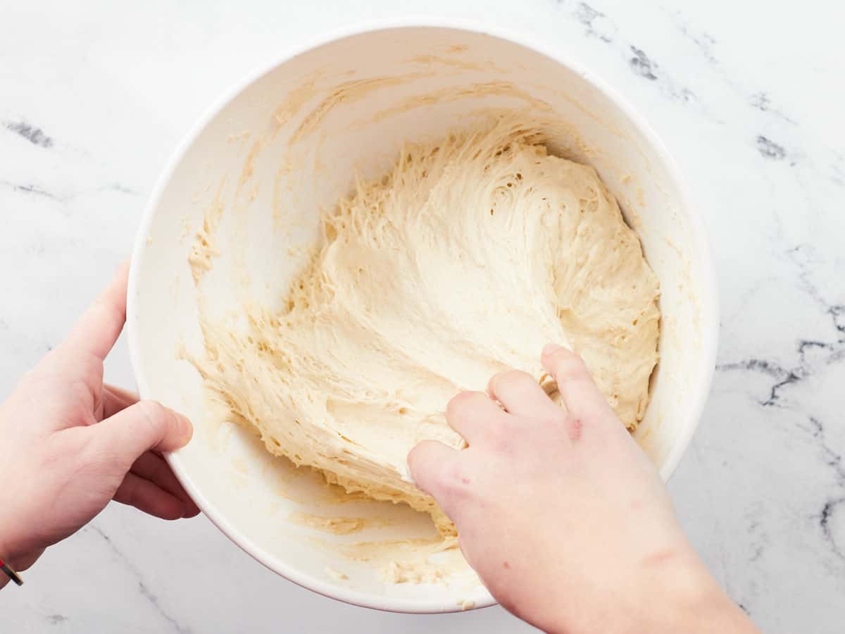 Ingredients for a no-knead focaccia style pizza crust after they have fermented for two hours in a white bowl. A left hand is stablizing the bowl with the right hand pulls the dough away from the side of the bowl.