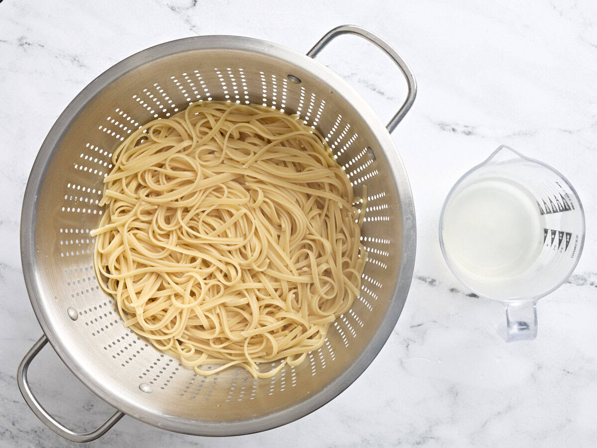 Boiled Fettuccine in a colander, next to a glass jug of reserved pasta water.