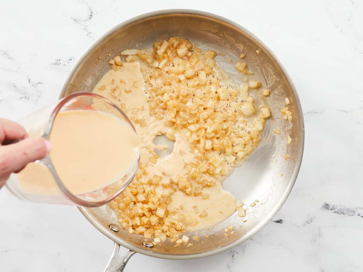 A vegetable stock and milk mixture being poured into a skillet with sauteed diced onions.