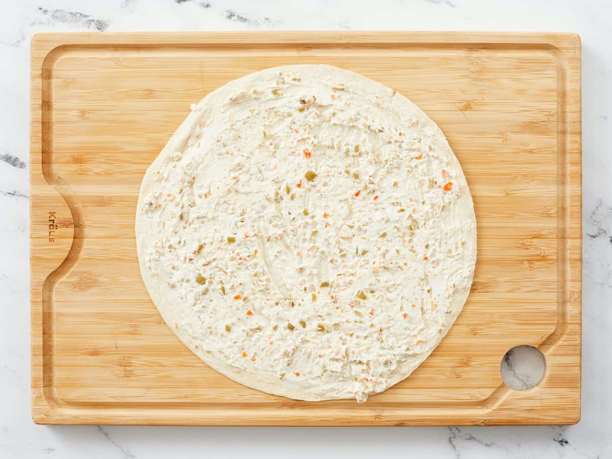 An extra large burrito shell smeared from edge to edge with a savory cream cheese spread, sitting on a wooden cutting board with a white marble background.