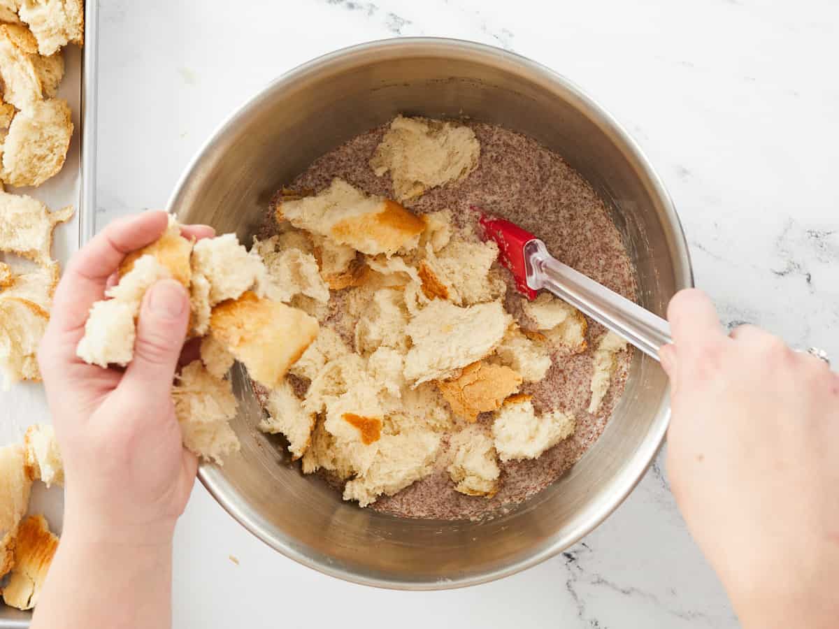 Hands adding pieces of torn bread into the left side of a large mixing bowl with the liquid ingredients for bread pudding while the right hand stirs with a red spatula. There is a tray of bread sitting in the upper left corner of the image, mostly out of frame.