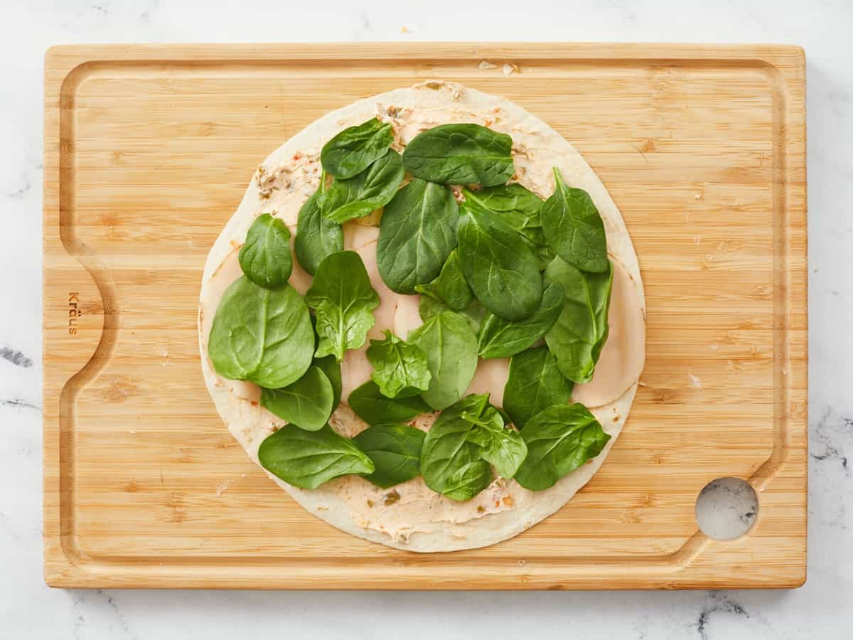A large tortilla on a wooden cutting board covered with savory cream cheese, a row of turkey slices and covered in a single layer of baby spinach leaves.