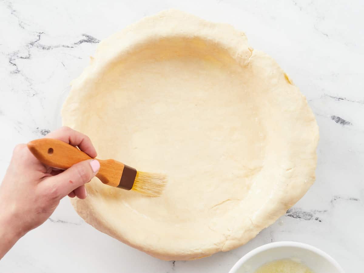 A hand using a brush to brush egg whites over the bottom of a pie crust.