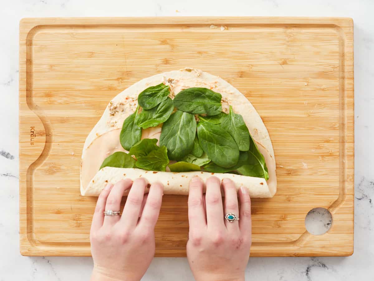 Hands rolling up a large tortilla on a wooden cutting board. The tortilla is covered with a savory cream cheese spread, a row of turkey slices and covered in a single layer of baby spinach leaves.