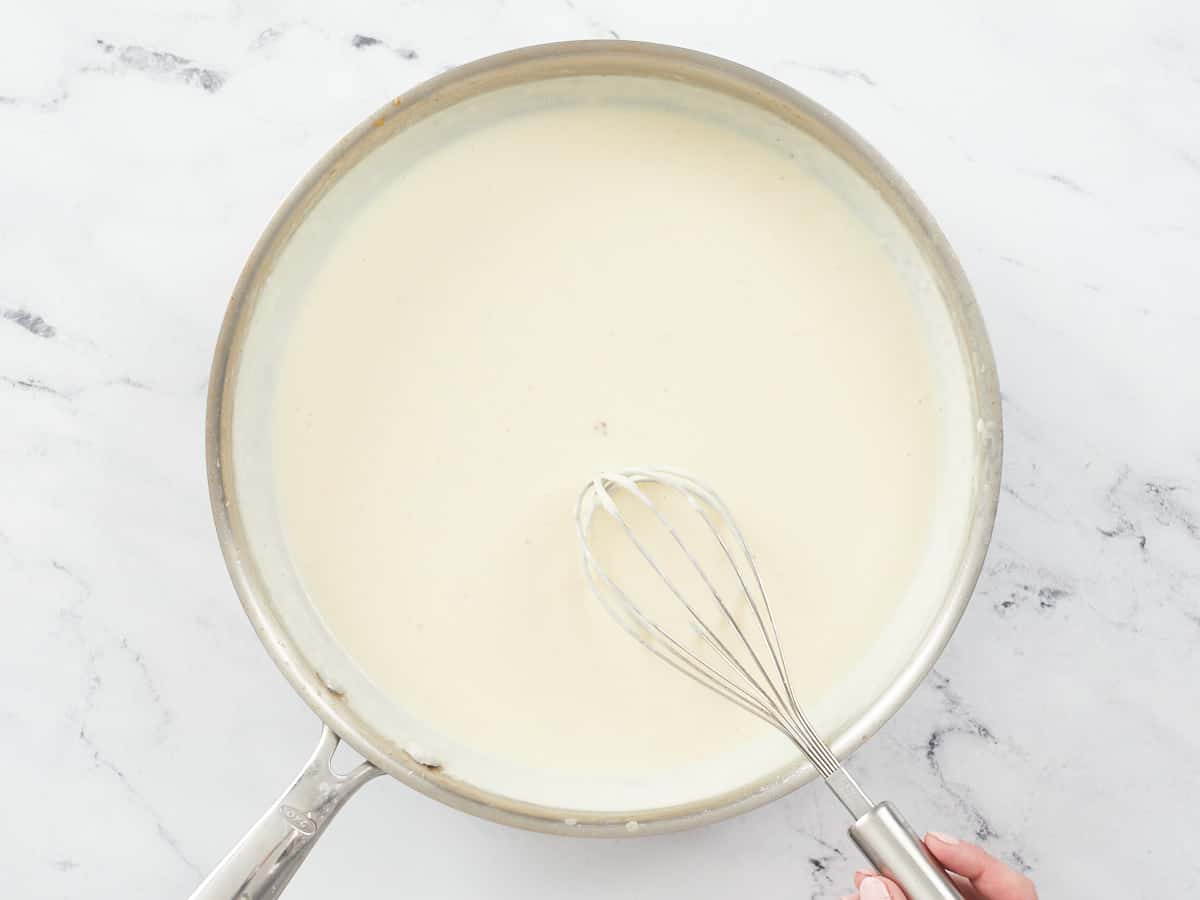 A large stainless steel skillet on a white marble background. The skillet is filled with a finished homemade alfredo sauce, and a hand in visible in the bottom right of the image and is stirring the mixture with a metal balloon whisk.
