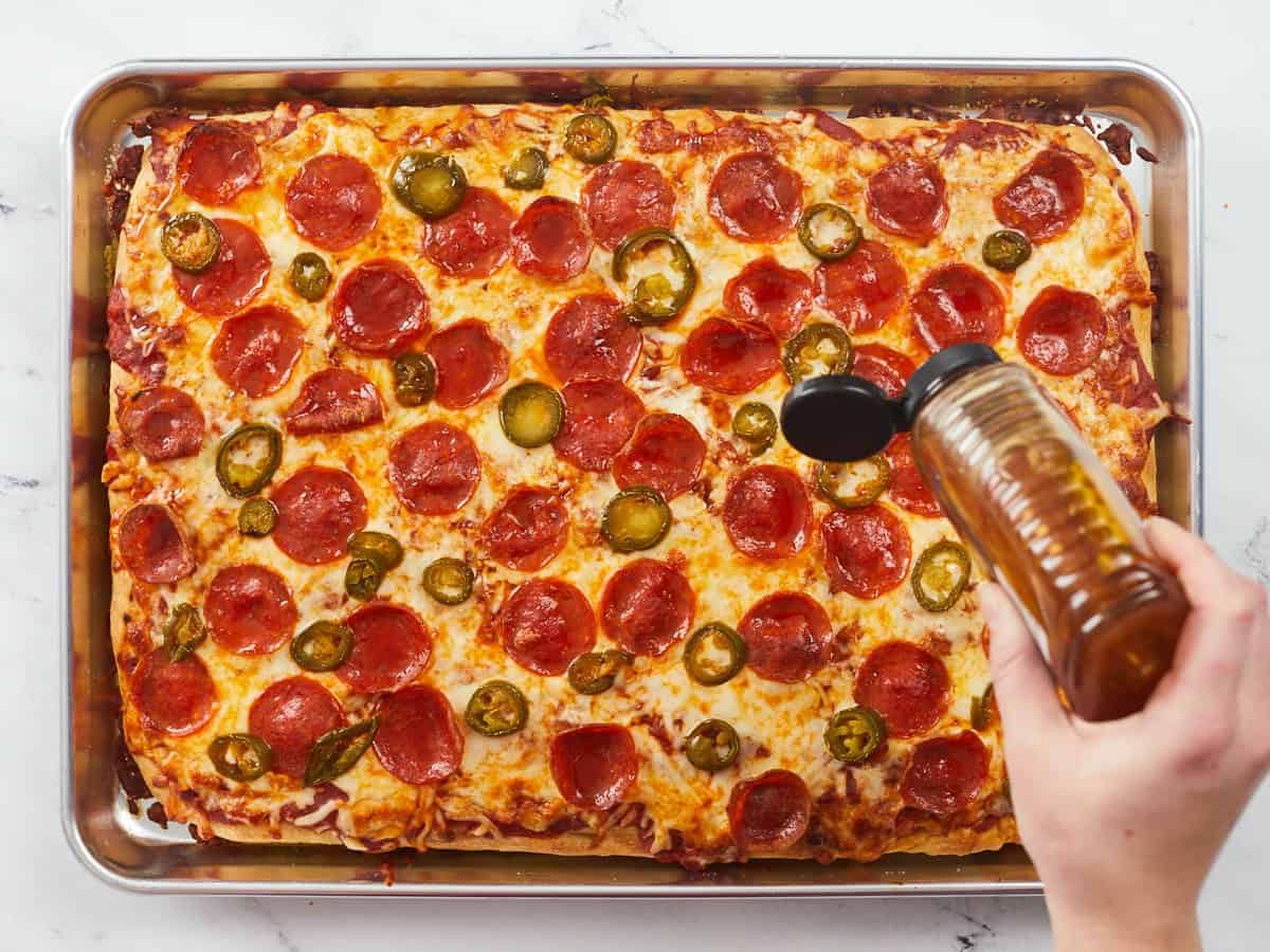 A fully baked sheet pan pizza that is golden brown and topped with crispy pepperonis, melted cheese and pickled jalapeño slices and a hand is drizzling honey on the right center of the pizza.