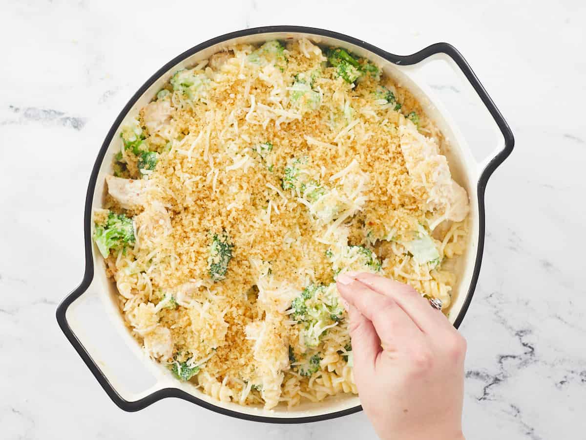 A round, white enamel cast iron baking dish with a black rim is sitting on a white marble background. The baking dish is filled with a mixture of cooked pasta, broccoli florets, cooked chunks of chicken breast and alfredo sauce. A hand in the lower right corner of the casserole dish is sprinkling toasted breadcrumbs over the pasta bake.