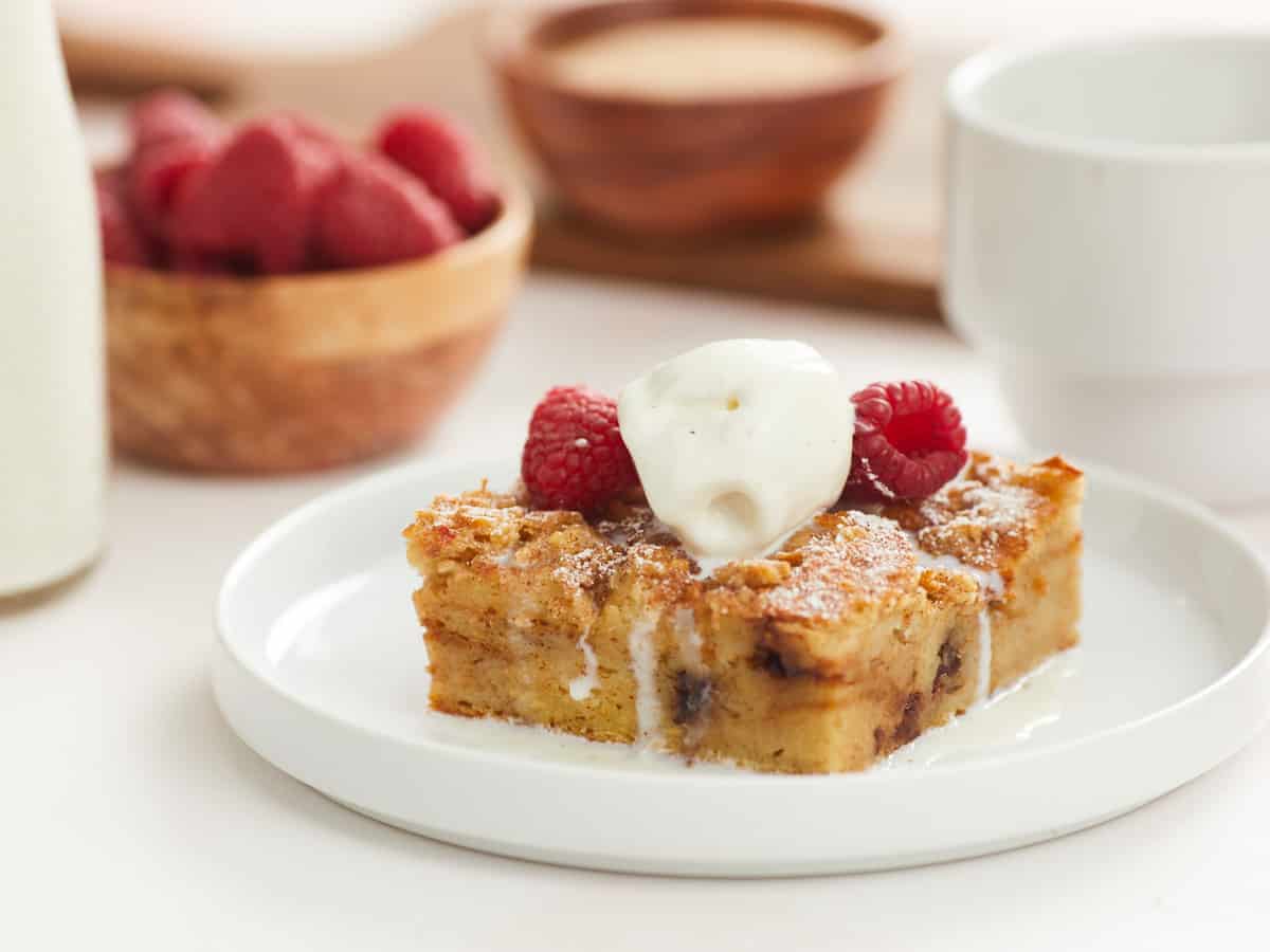 A horizontal image of a single serving of bread pudding on a flat white plate topped with two raspberries and a scoop of vanilla ice cream. In the background, there are blurred bowls of toppings ingredients in the recipe, like a small bottle of heavy cream, a bowl of fresh raspberries and a bowl of cinnamon sugar topping.