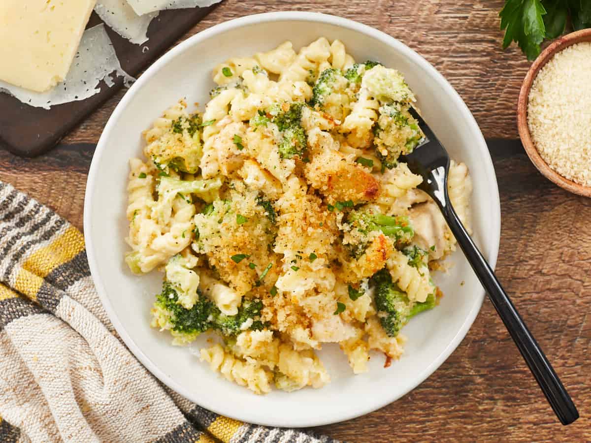 A large serving of chicken alfredo bake in a flat, white bowl, and a black fork is resting on the right side of the bowl. The bowl is sitting on a wood background surround with a yellow and gray striped dish cloth, a dark wooden cutting board with Parmesan Cheese shavings on it, and a sprig of parsley and a bowl of untoasted breadcrumbs.