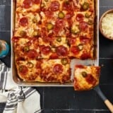 A vertical image of a baked sheet pan pizza topped with pizza sauce, cheese, pepperonis, pickled jalepenos and honey sliced and a spatula is removing the bottom right slice.