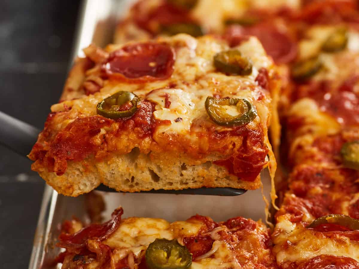 An extreme close up of a single slice of sheet pan pizza raised above the rest of the pizza with a spatula.