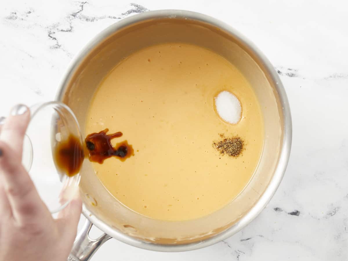 A hand is pouring Worcestershire sauce into a small saucepot of melted cheese, and there are small piles of unmixed salt and black pepper sitting on the surface.