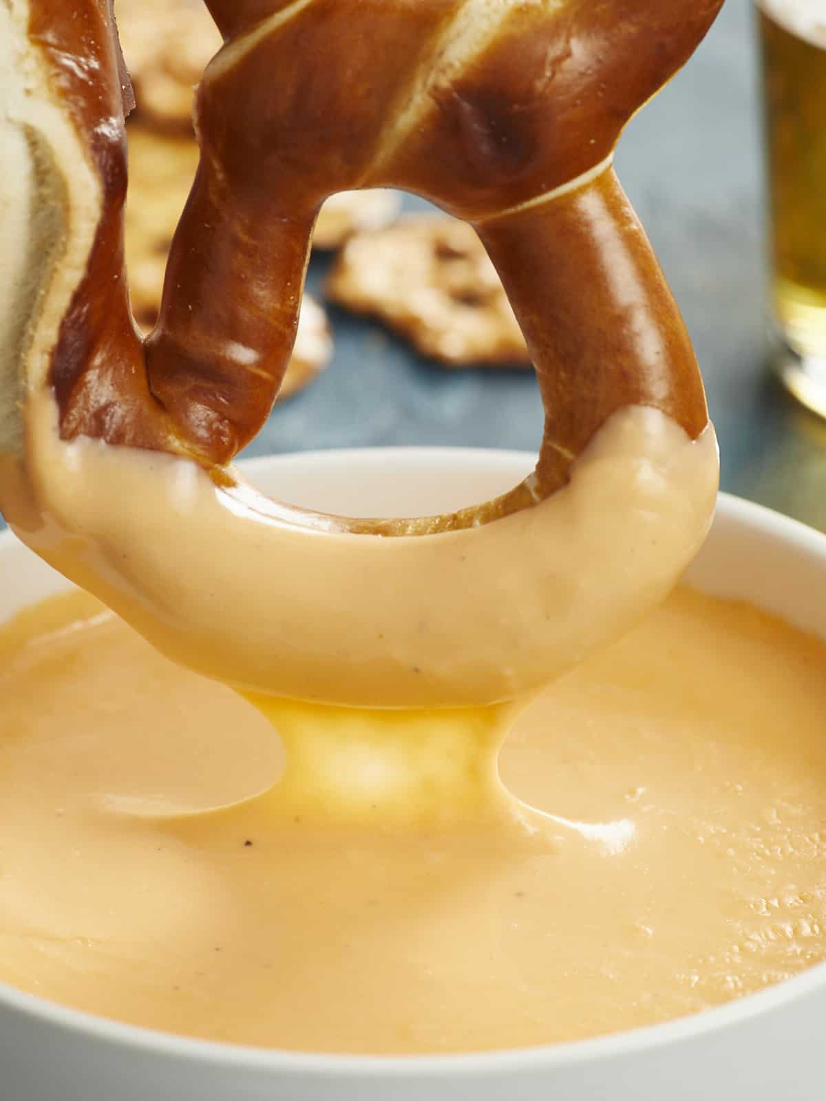An extreme close up of a giant Bavarian soft pretzel that's just been dipped into a white bowl filled with melted cheese sauce, and an assortment of pretzels and a glass cup of beer are blurred in the background.