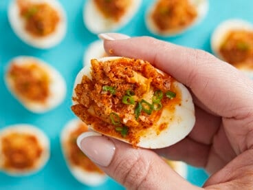 Close up of hand holding a Nashville Hot Deviled Egg with other eggs in the background on an aqua backdrop.