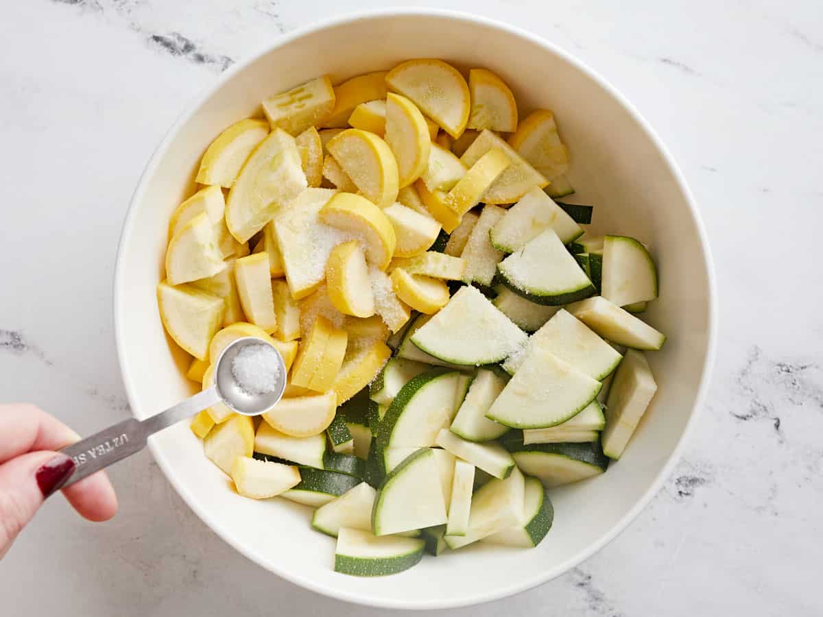 Sliced zucchini and squash in a bowl with salt being sprinkled over top.