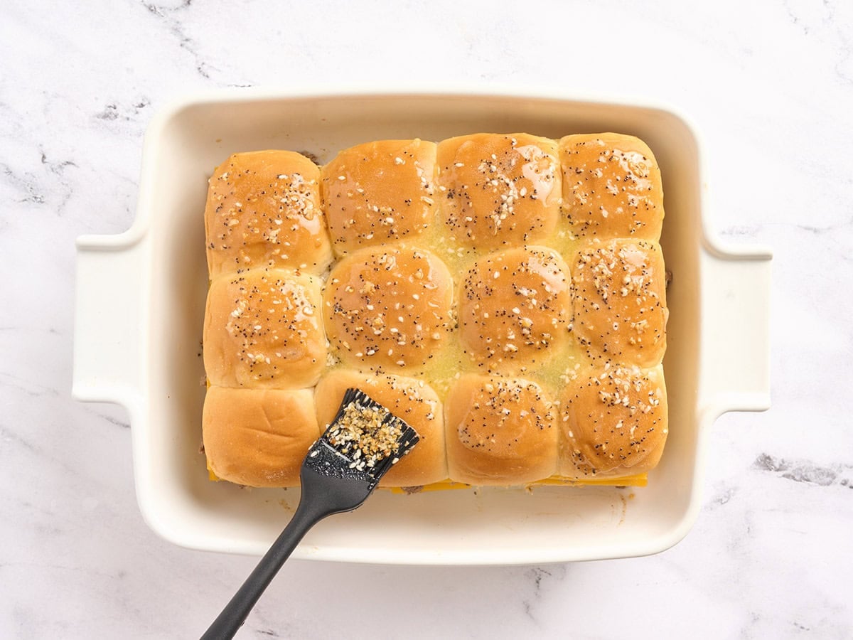 A pastry brush brushing seasoned melted butter over sliders in a casserole dish.