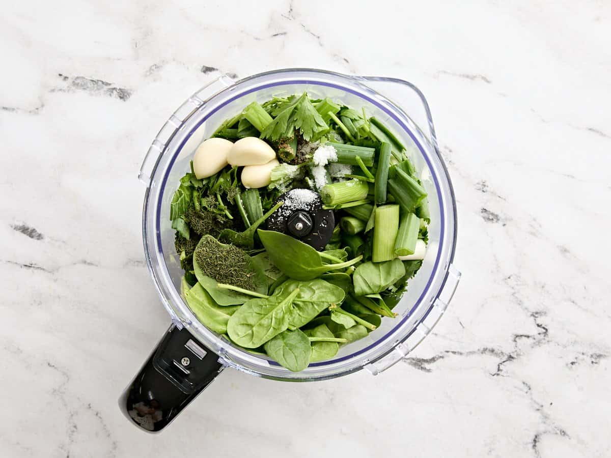 Fresh herbs, garlic cloves, salt, and green onions in a food processor.