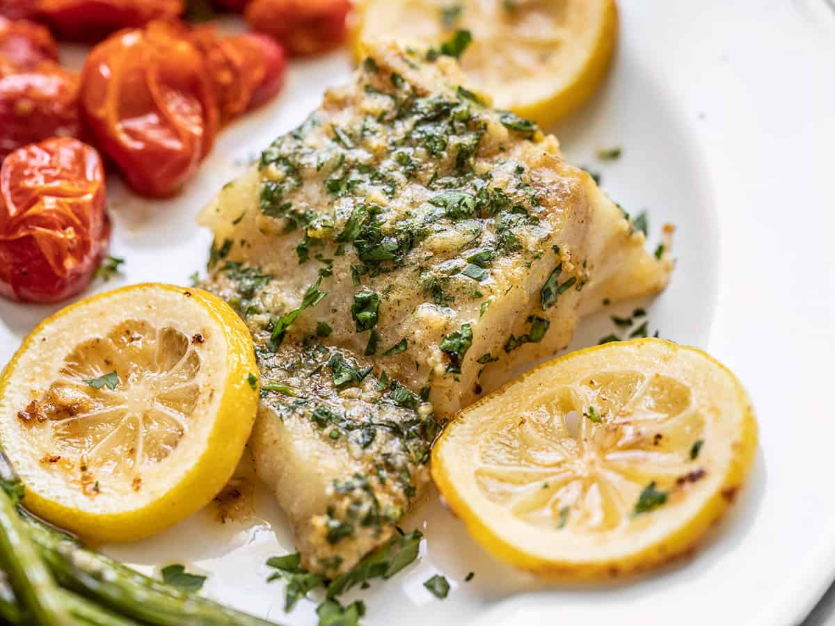 This rich and zesty Garlic Butter Baked Cod is a fast and flavorful weeknight dinner that can be made in under 30 minutes, using basic pantry staples. BudgetBytes.com Close up side view of a piece of garlic butter baked cod on a serving plate with asparagus and tomatoes.