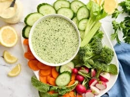 Overhead view of a bowl of green goddess dressing with a plate of fresh cut vegetables.