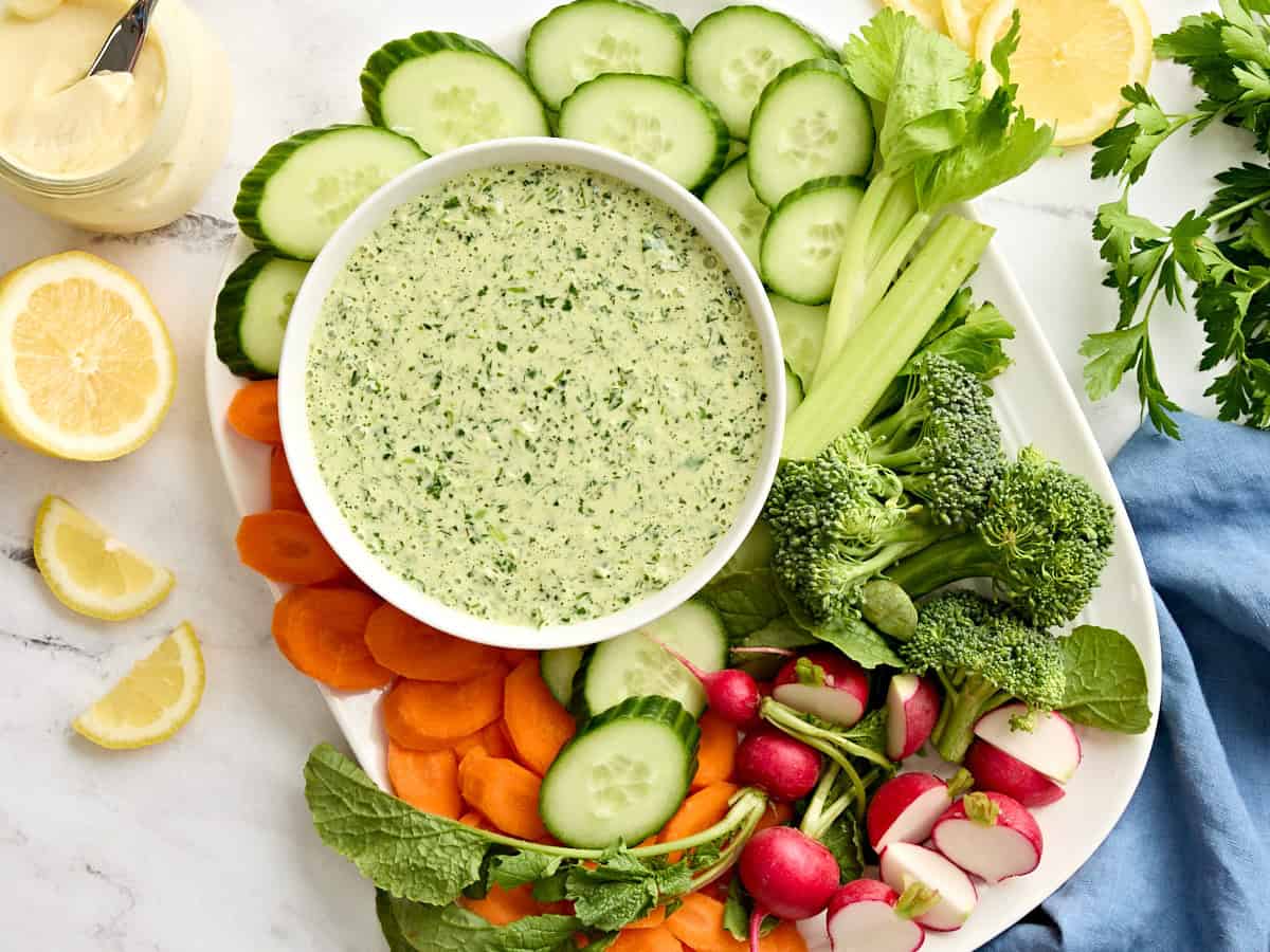 Overhead view of a bowl of green goddess dressing with a plate of fresh cut vegetables.