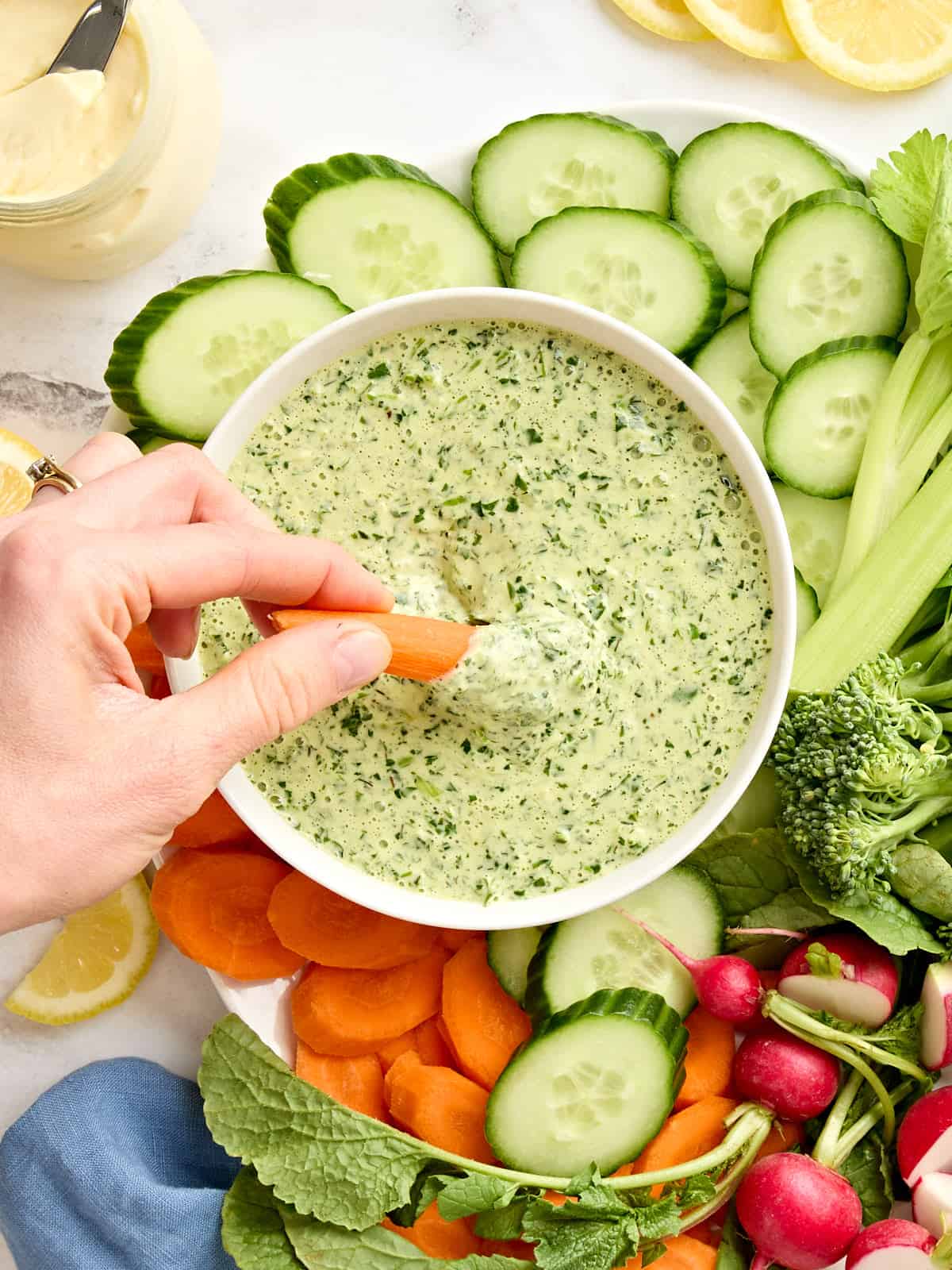 Overhead view of a bowl of green goddess dressing on a plate of fresh cut vegetables, with a hand dipping a carrot in.