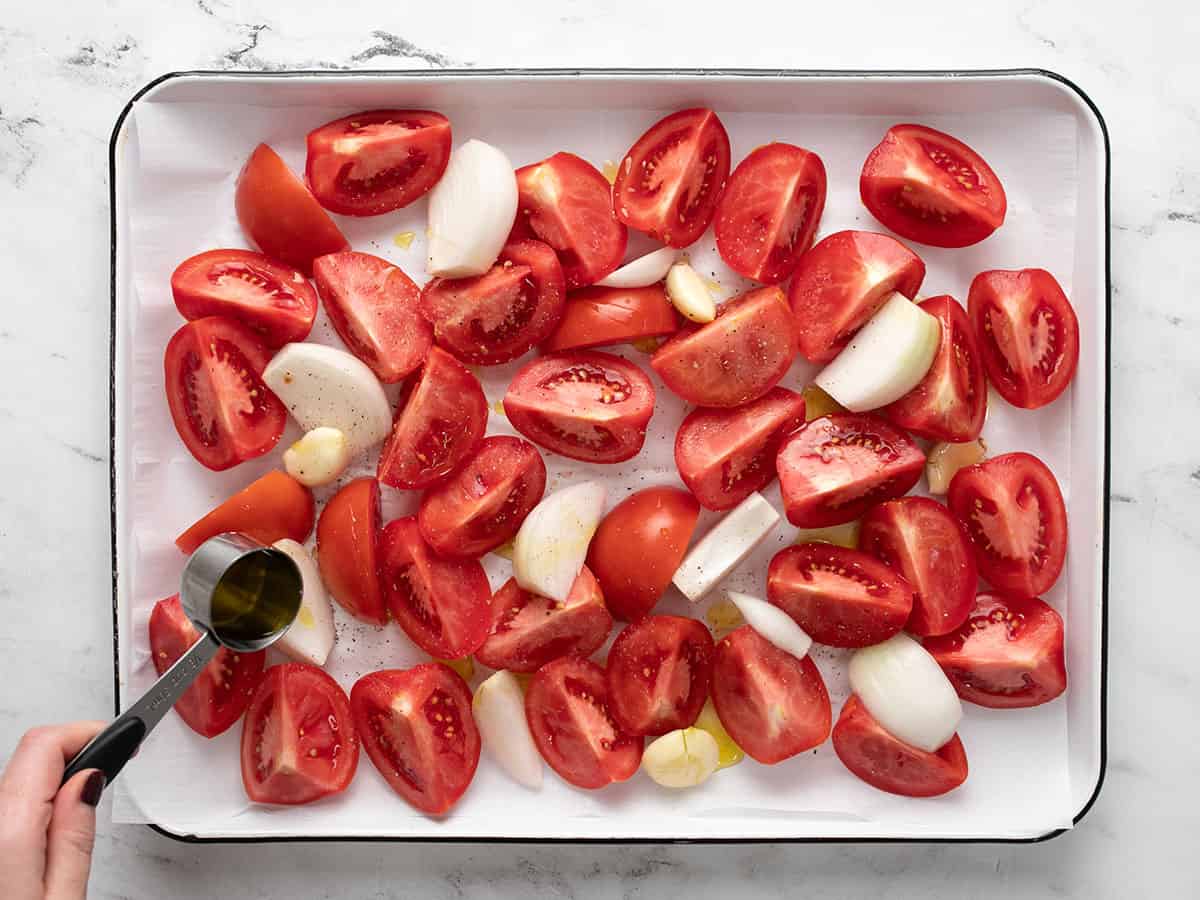 Tomatoes, onions, and garlic on a sheet pan being drizzled with olive oil.