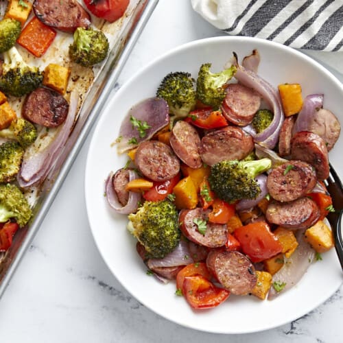 A serving of Sheet Pan Chicken Sausage and roasted vegetables on a white plate next to a sheet pan.